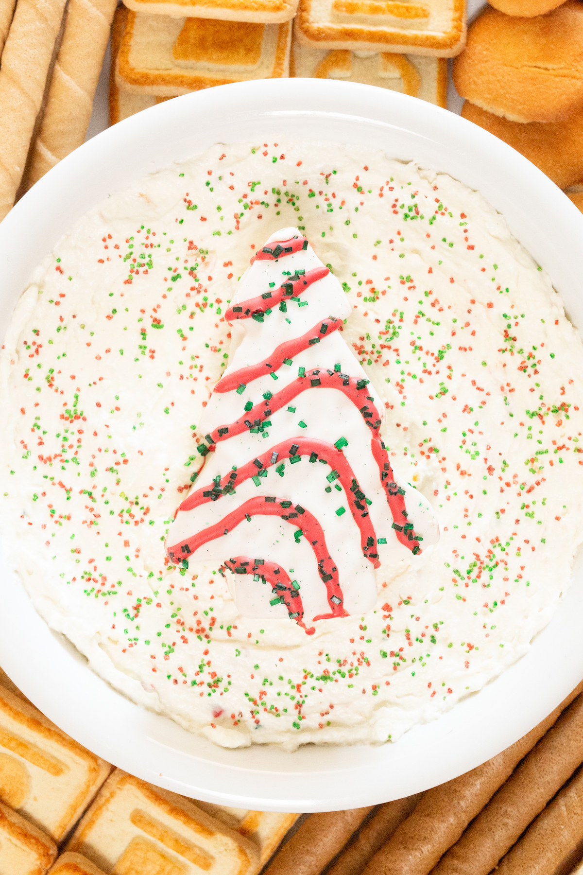 A white serving bowl filled with white Christmas Tree Cake Dip. Garnished with red and white sprinkles and a Christmas tree cake.