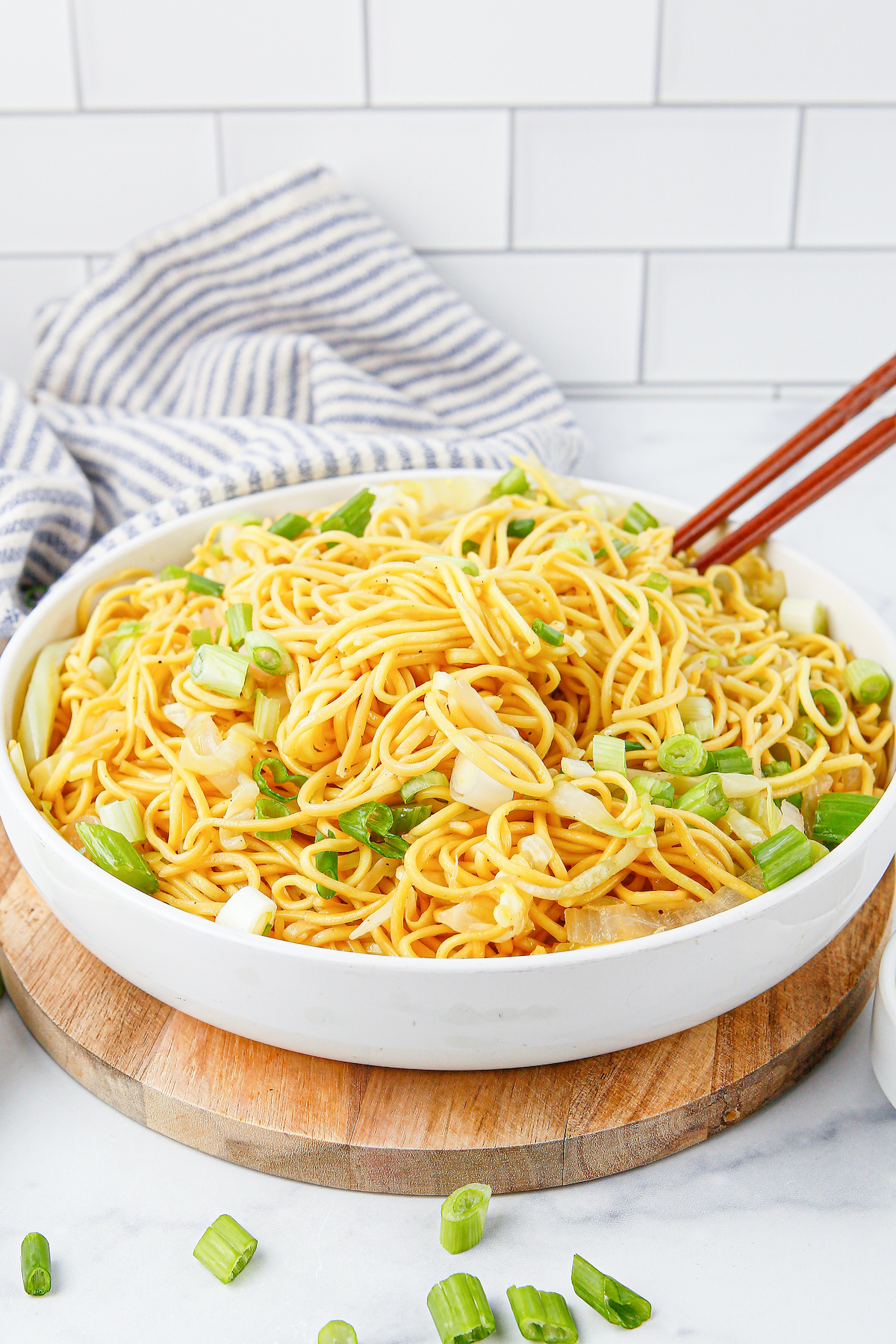 A shallow white serving bowl full of chow mein noodles that are garnished with sliced green onion and chopsticks.