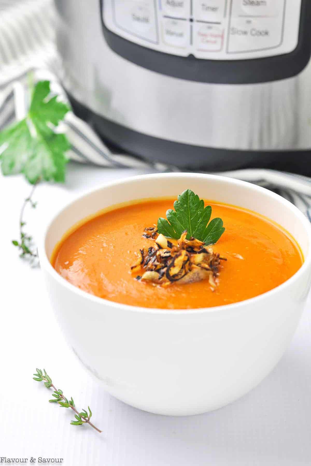 A white soup bowl filled with bright orange carrot & ginger soup that's sitting in front of an Instant Pot.