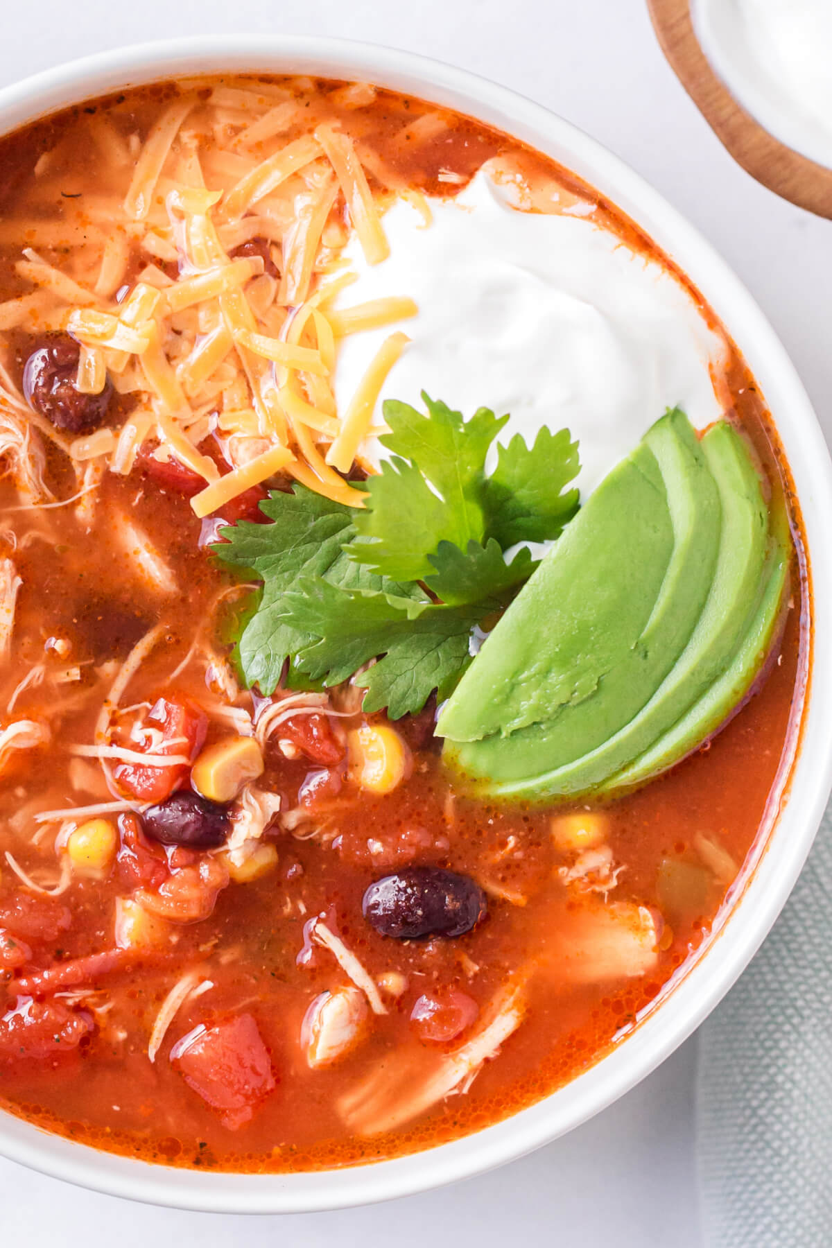 A white bowl filled a serving of Instant Pot enchilada soup that's garnished with avocado slices, sour cream, shredded cheese, and cilantro.