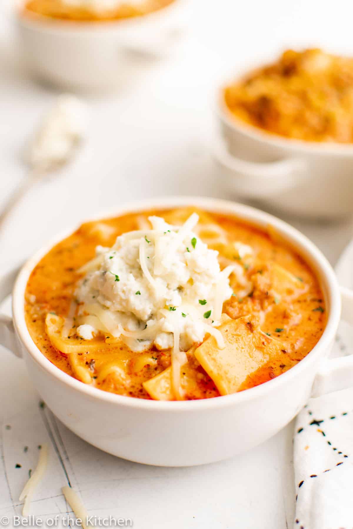 A white bowl filled with lasagna soup that's garnished with a dollop of ricotta and shredded cheese.