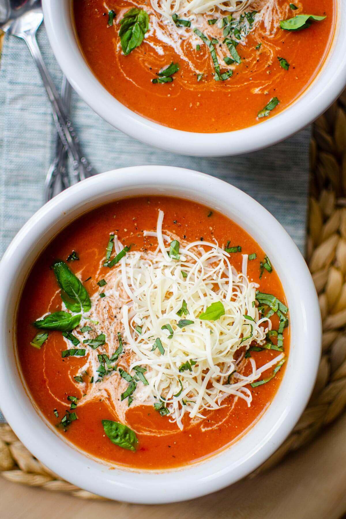 Overhead view of two white soup bowls filled with red tomato soup that's topped with shredded white cheese.