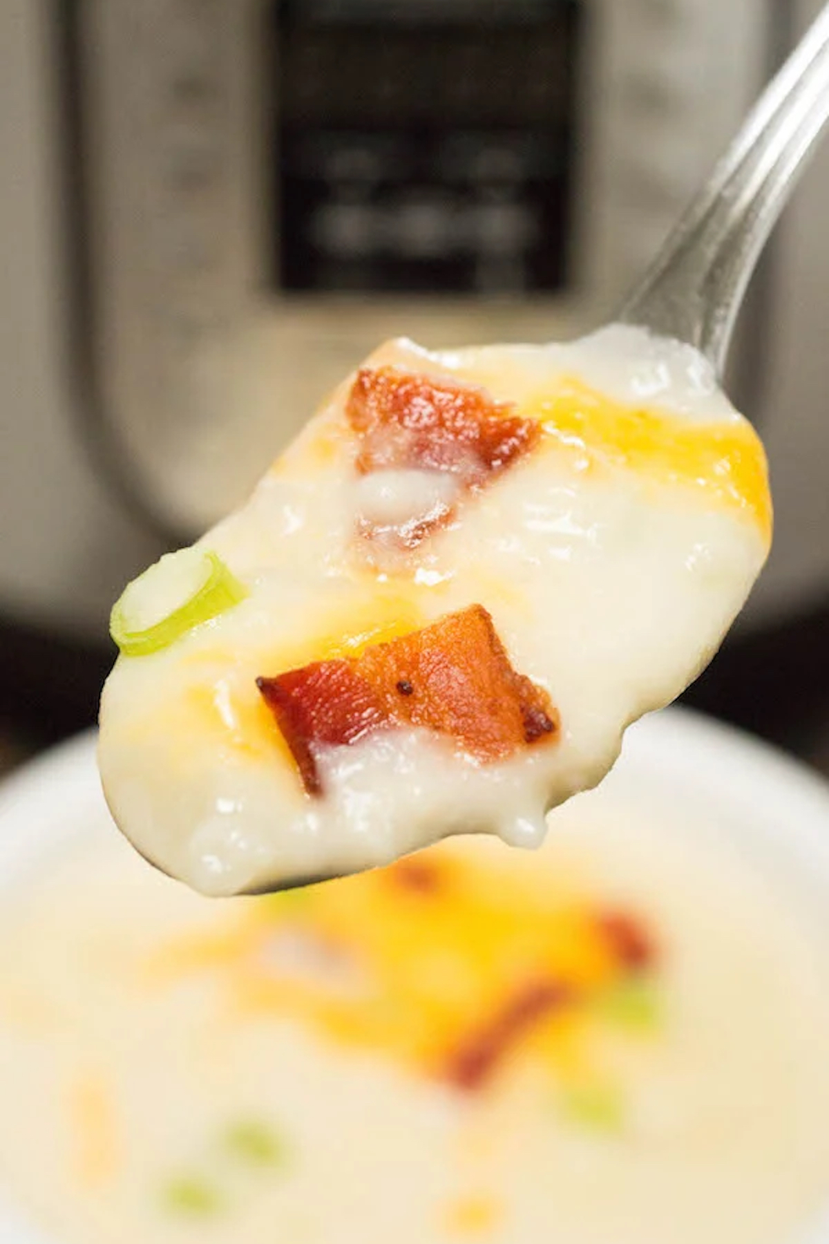 A spoonful of Instant Pot loaded baked potato soup is in focus, the appliance and bowl of the soup is out of focus in the background.