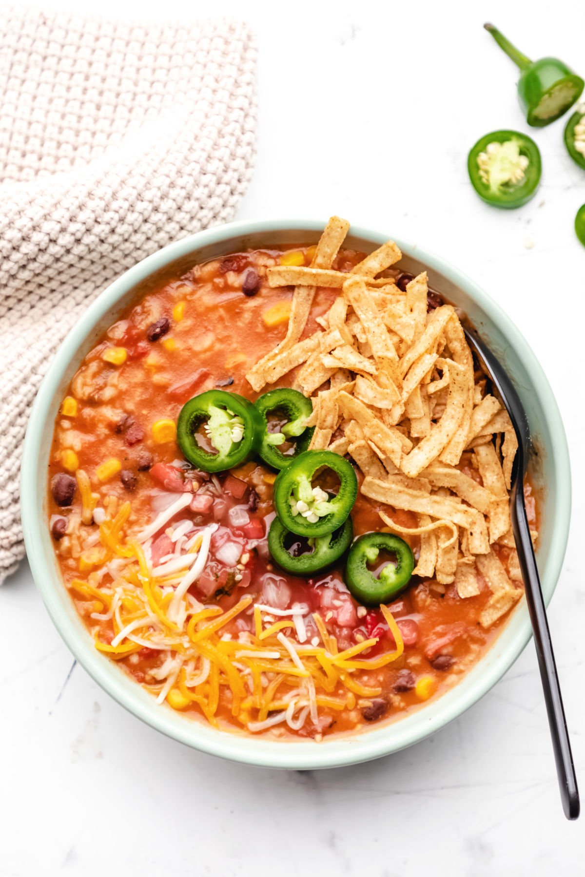 A soup bowl filled with vegetarian tortilla soup that's garnished with shredded cheese, sliced jalapeños, and tortilla strips.
