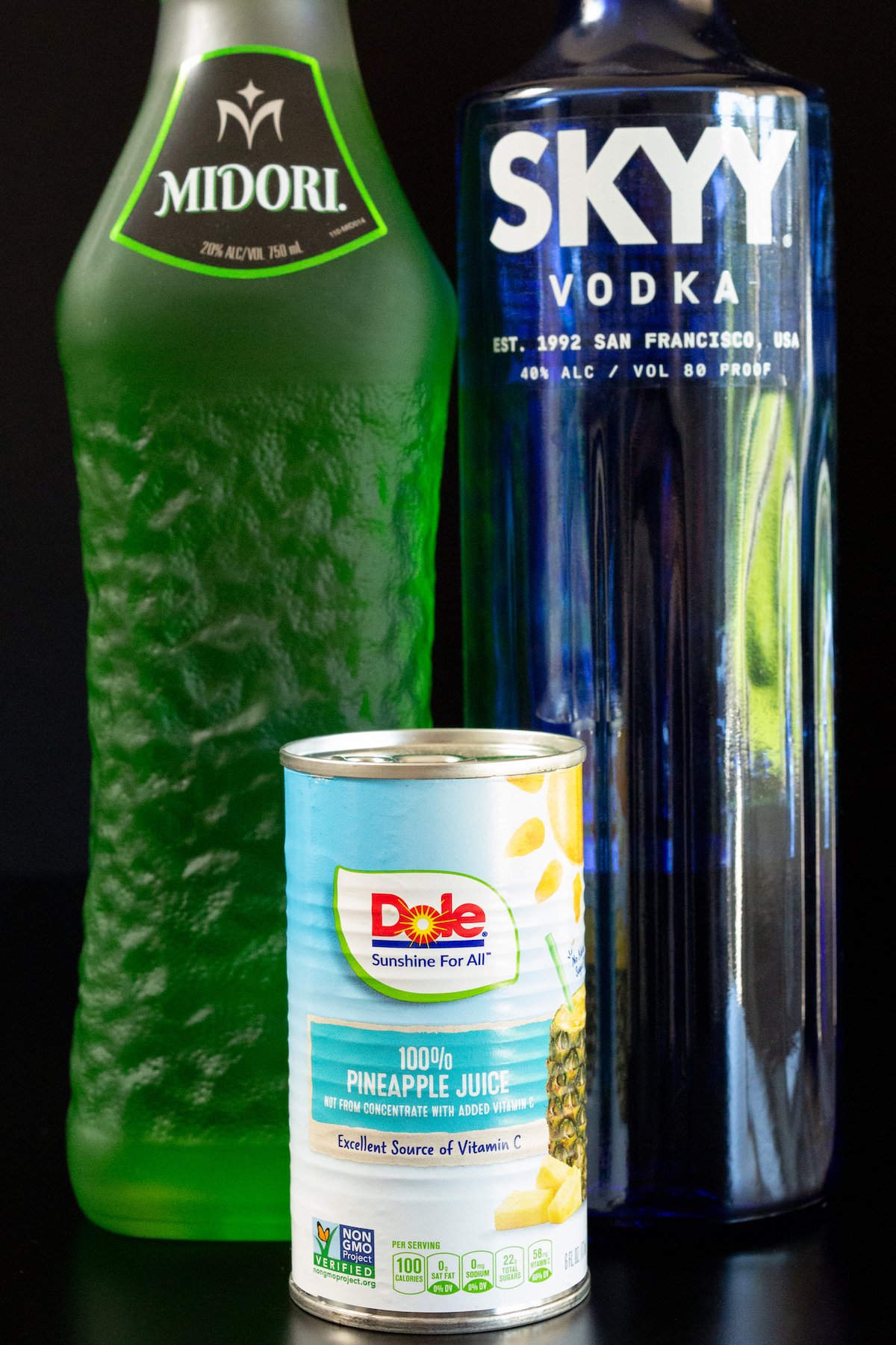 A bottle of Midori, Skyy Vodka, and a can of pineapple juice on a black background.