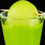 A single shot glass filled with a bright green melon ball shot, garnished with a honeydew melon ball on a toothpick.