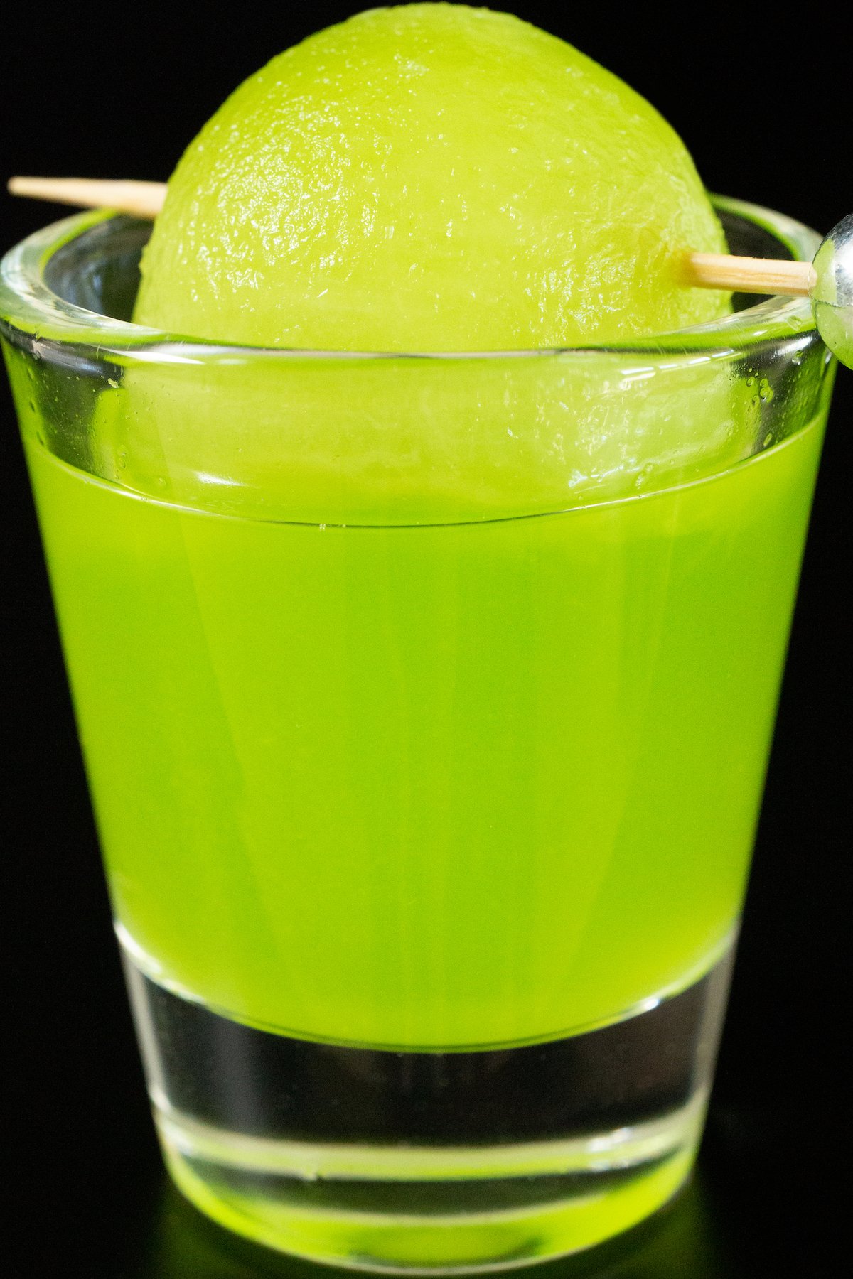A single shot glass filled with a bright green melon ball shot, garnished with a honeydew melon ball on a toothpick.
