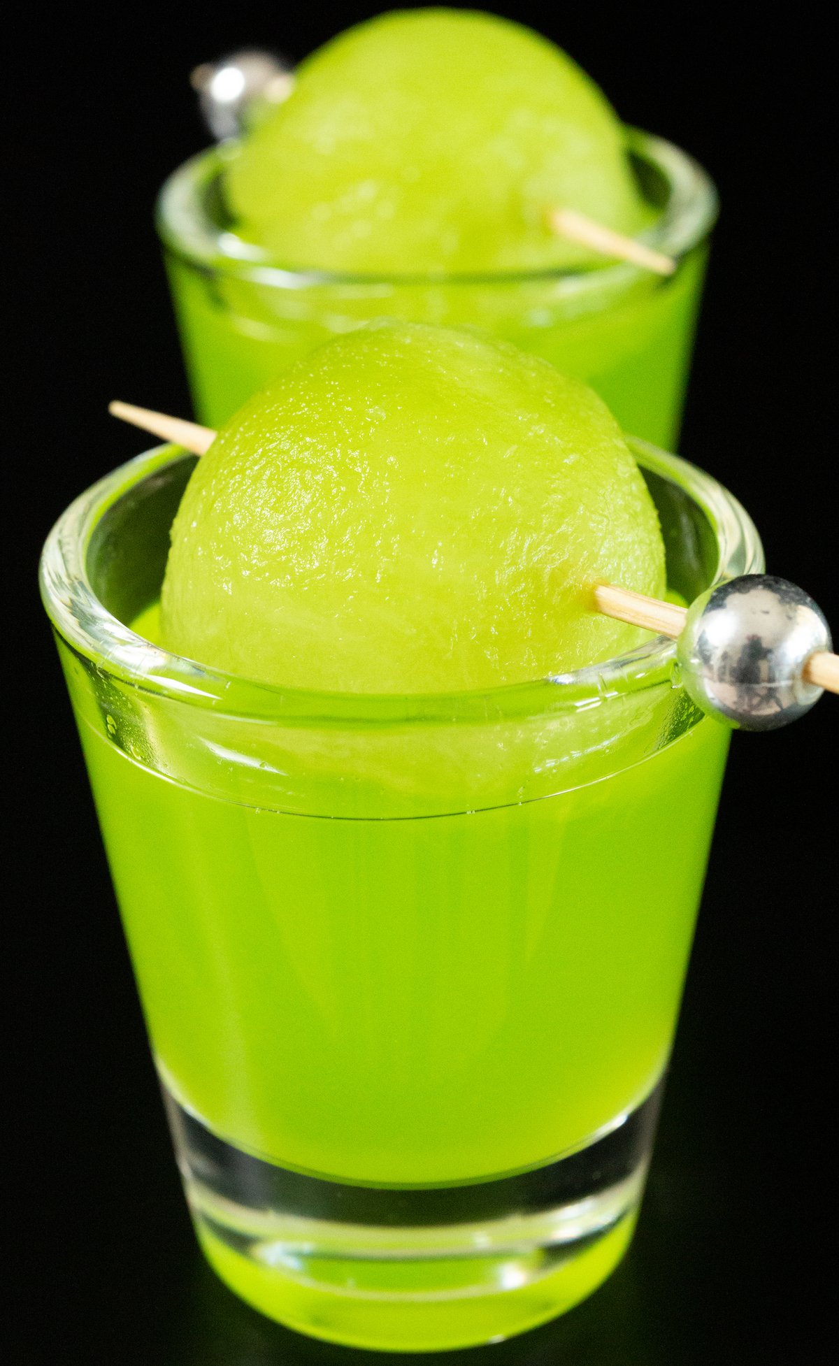 Two shot glasses filled with bright green melon ball shots on a black background.
