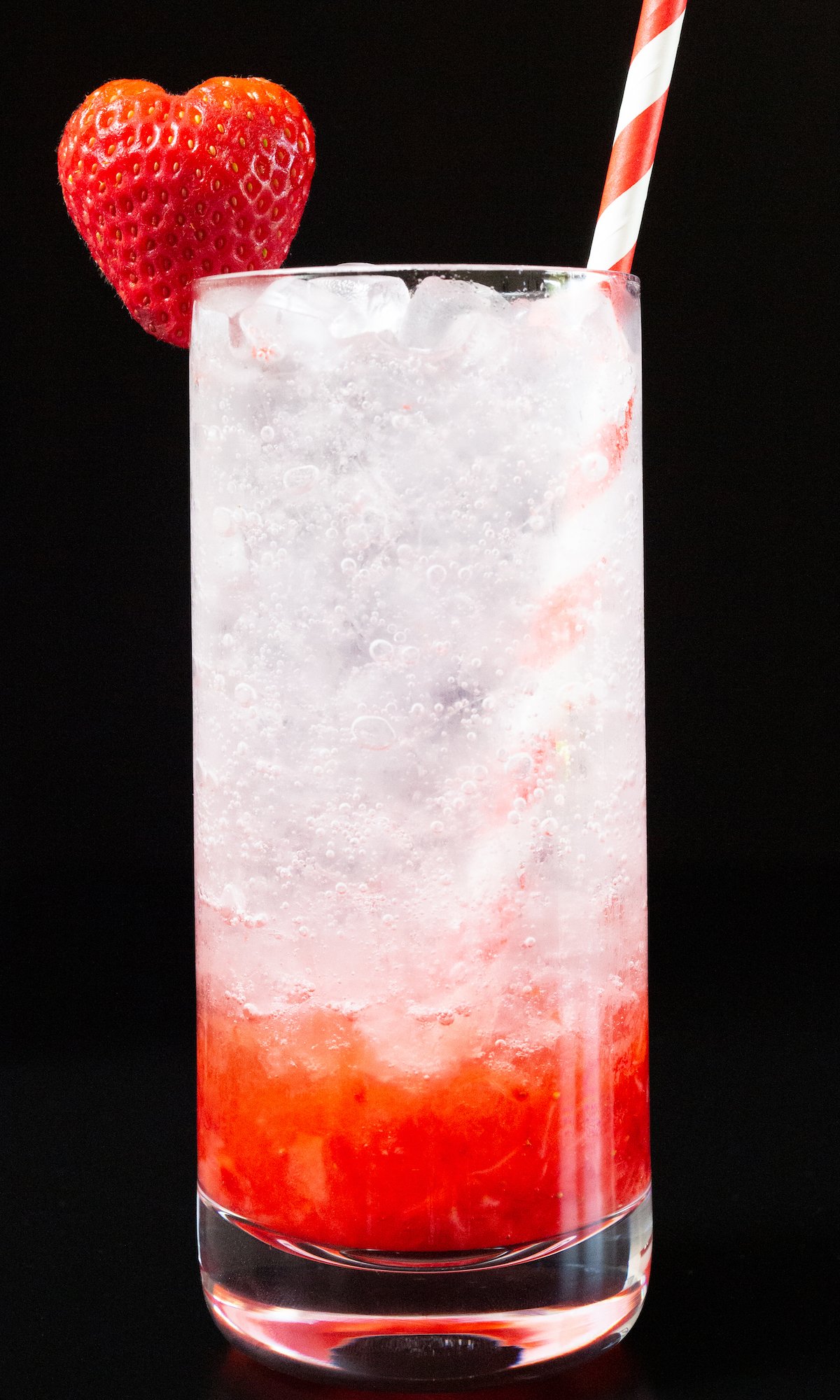 A highball glass is filled with a strawberry gin cocktail, which has red strawberry puree on the bottom and white club soda on top. Garnished with a heart shaped strawberry & red and white striped straw.
