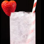 A highball glass is filled with a strawberry gin cocktail, which has red strawberry puree on the bottom and white club soda on top. Garnished with a heart shaped strawberry & red and white striped straw.