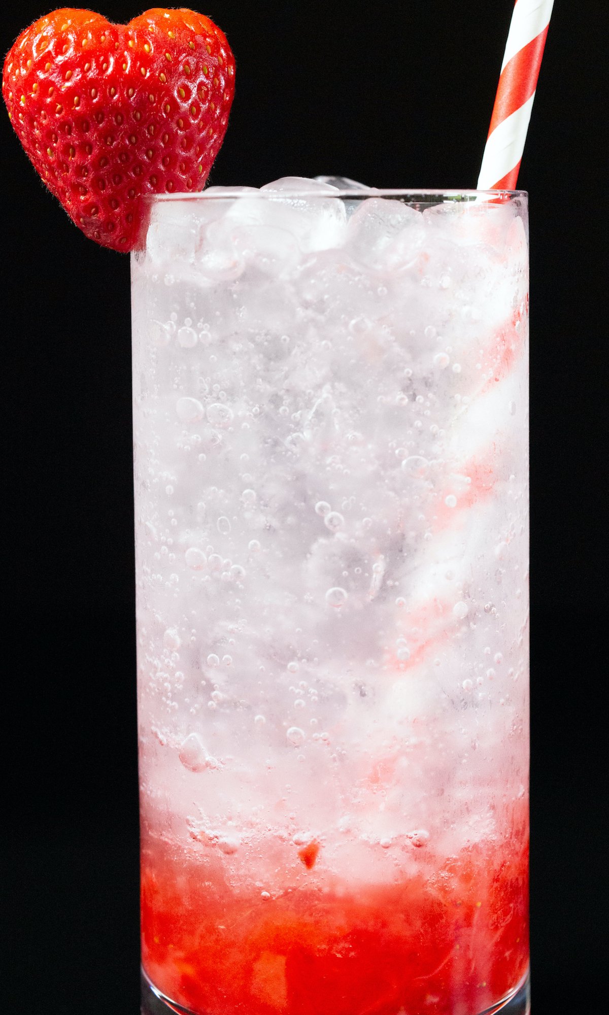 A highball glass is filled with a strawberry gin cocktail, which has red strawberry puree on the bottom and white club soda on top. Garnished with a heart shaped strawberry & red and white striped straw.