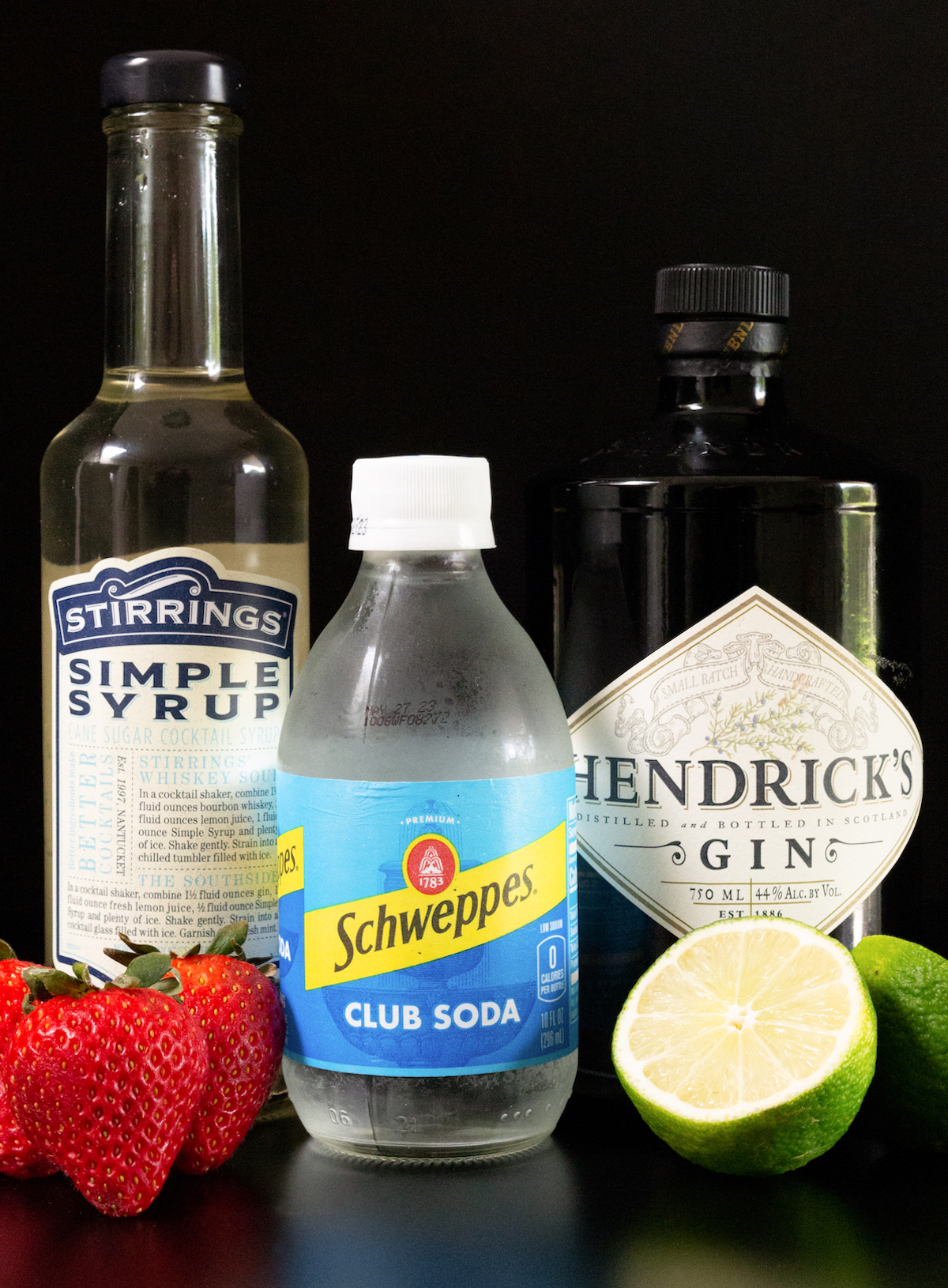 A bottle of simple syrup, gin, cub soda, fresh strawberries, and a lime sliced in half on a black background.
