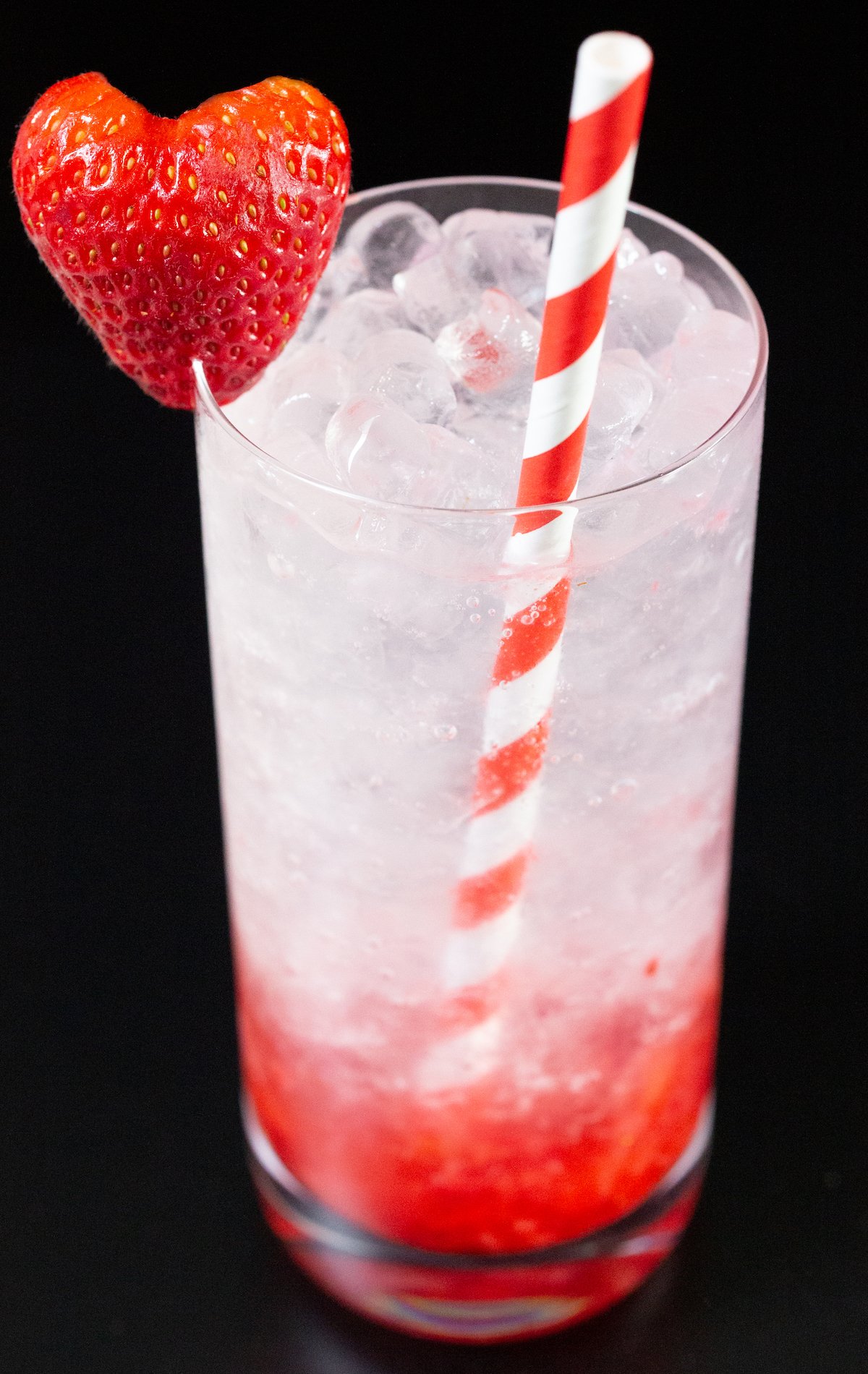 A highball glass filled with a strawberry gin smash that has muddles strawberries on the bottom and cum soda and pebble ice in the top part of the glass. Heart shaped strawberry and red & white paper straw garnish.