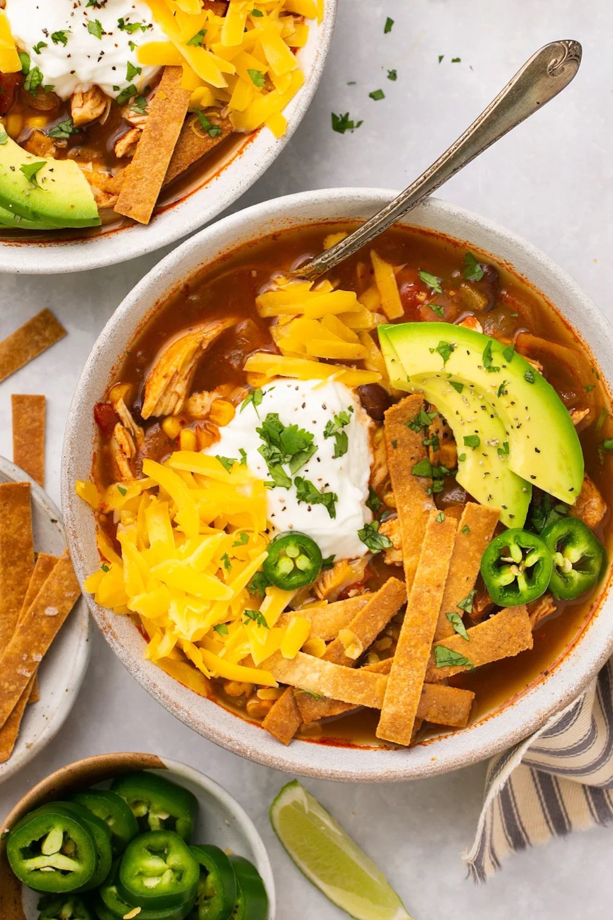 A bowl of copycat Chik-fil-a tortilla soup in a ceramic bowl. Topped with shredded cheddar cheese, sliced jalapeno and avocado, sour cream, and tortilla strips.