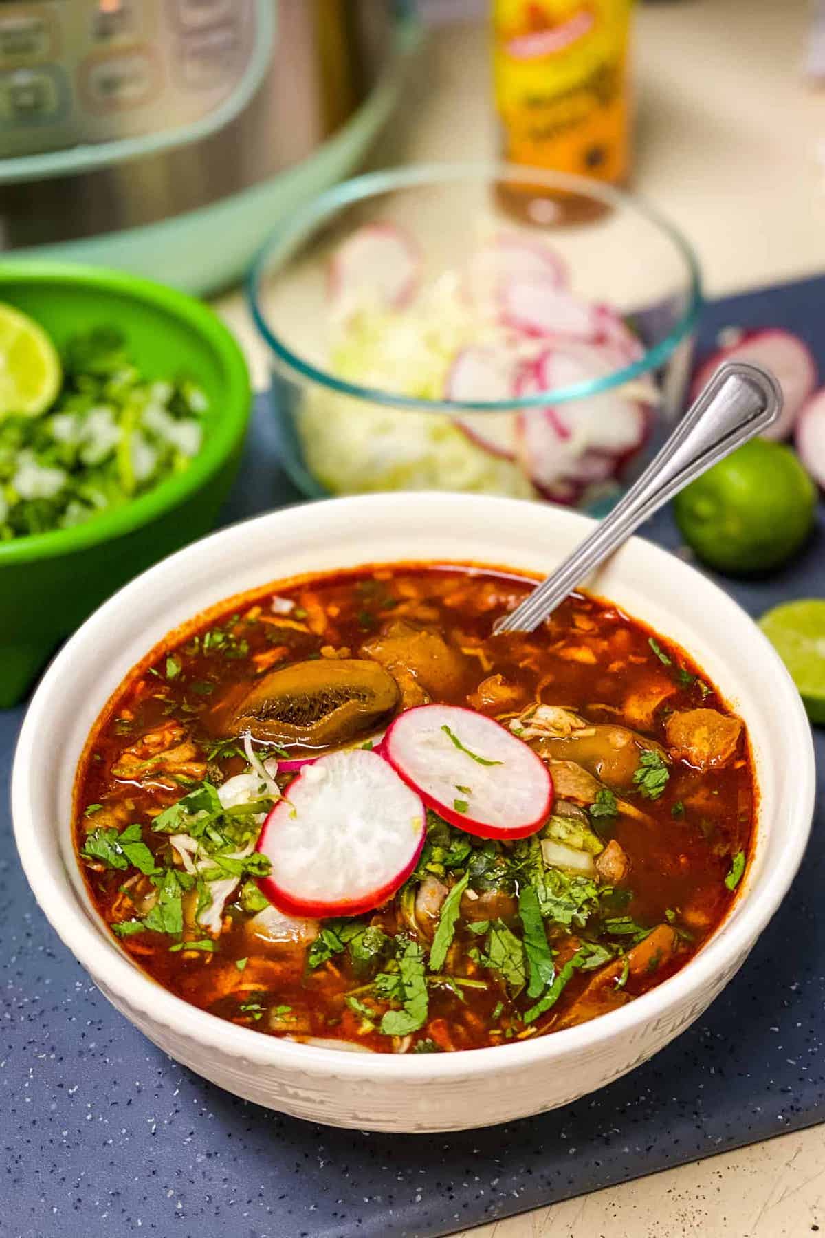 A bowl of vegan Instant Pot Pozole garnished with sliced radishes and fresh herbs in front of glass prep bowls of ingredients.