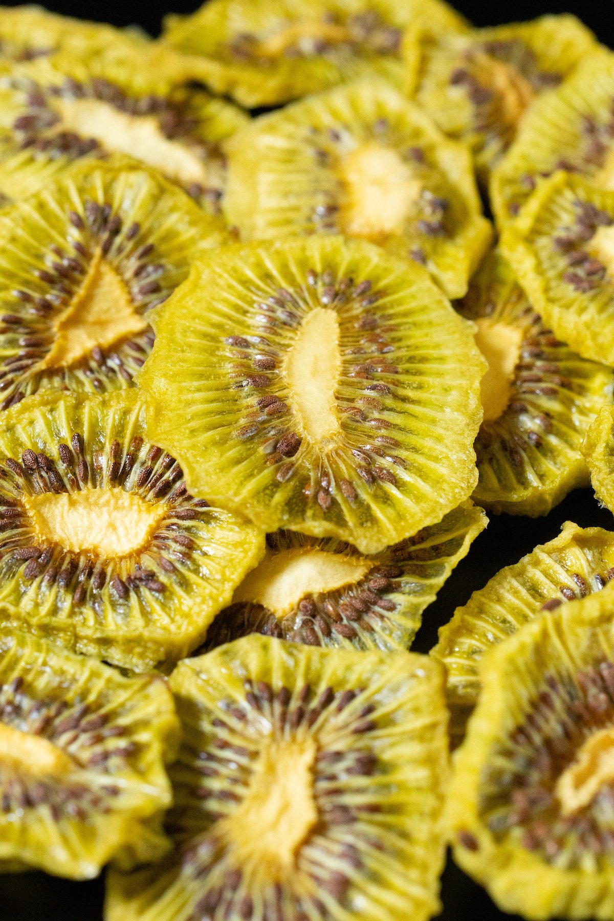 Dozens of dehydrated kiwi chips in a pile on a black background.