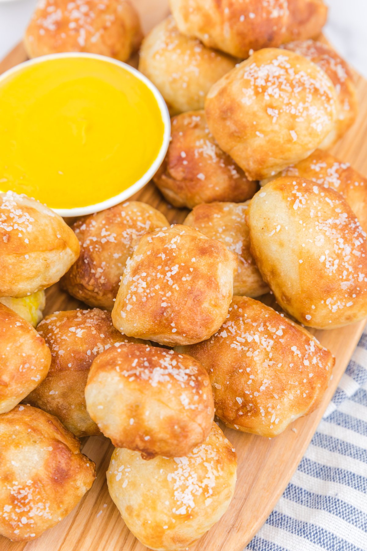 A cutting board with a few dozen air fryer pretzel bites stacked on it next to a small bowl of mustard.