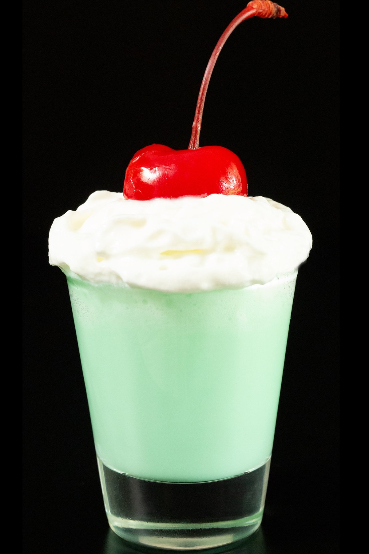 A single shot glass on a white background is filled with mint colored shamrock shot mixture that's topped with whipped cream and a maraschino cherry.