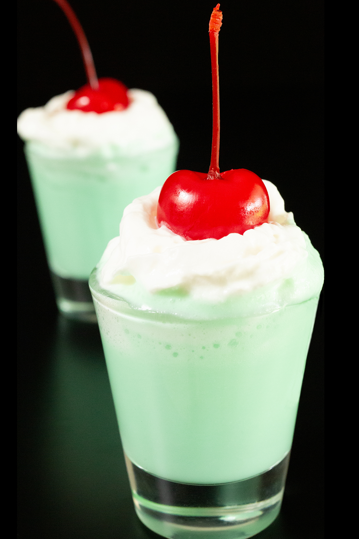 Two shot glasses filled with mint green colored Shamrock Shots are topped with whipped cream and maraschino cherries.