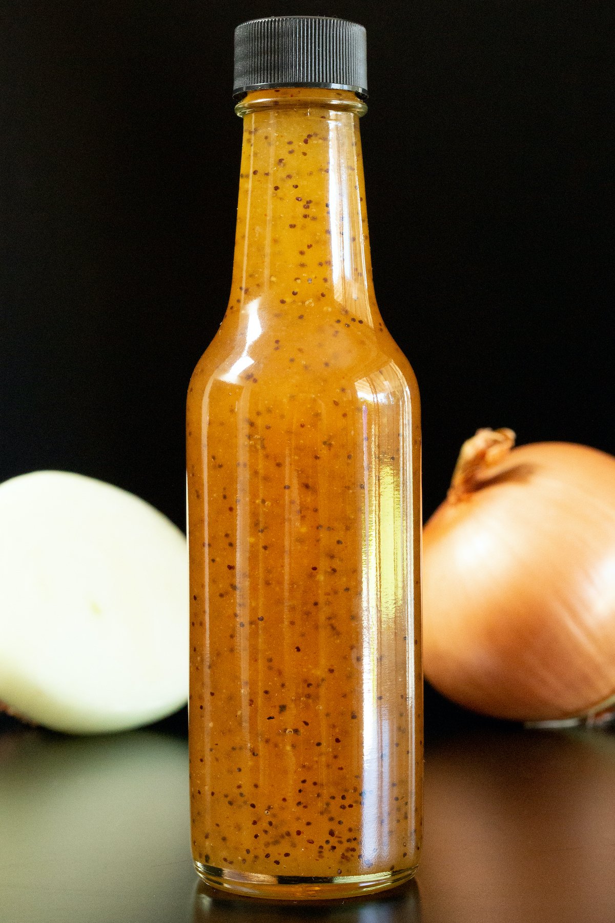 A small glass bottle filled with brown copycat subway sweet onion sauce is in focus. A sliced and unsliced onion are out of focus in the background.