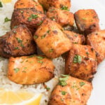 Bite sized pieces of salmon cooked in the air fryer on a white dinner plate with rice and lemon slices.