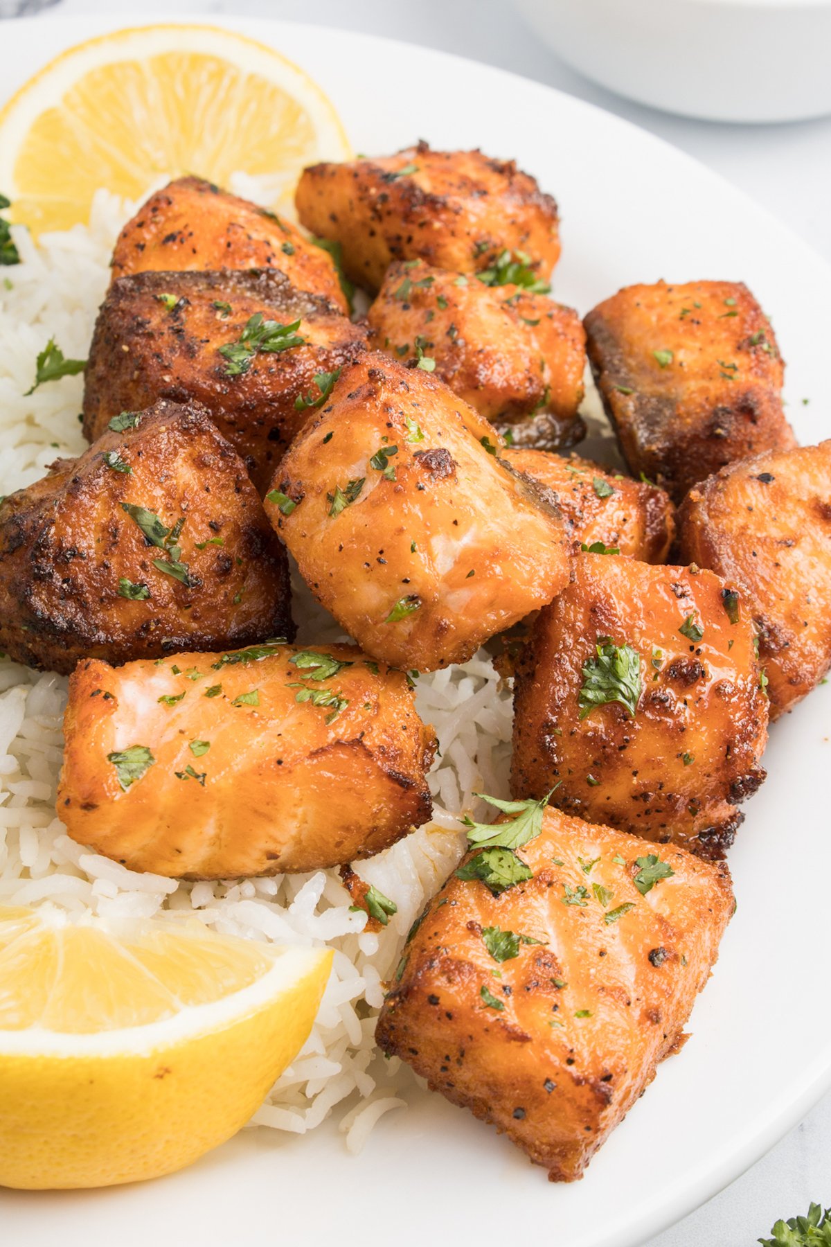 Bite sized pieces of salmon cooked in the air fryer on a white dinner plate with rice and lemon slices. 