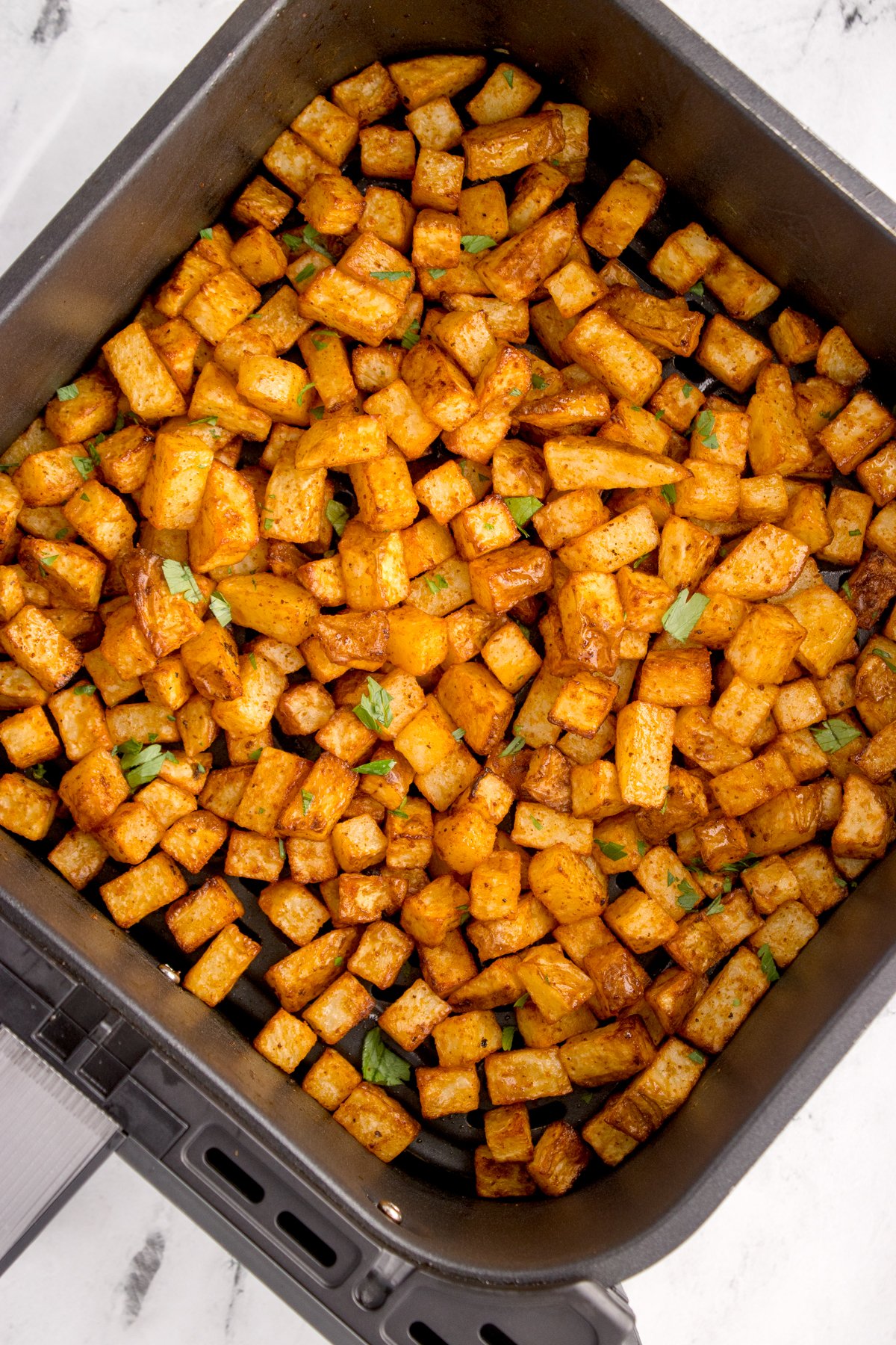 An air fryer basket filled with cooked diced breakfast potatoes that are garnished with chopped parsley.