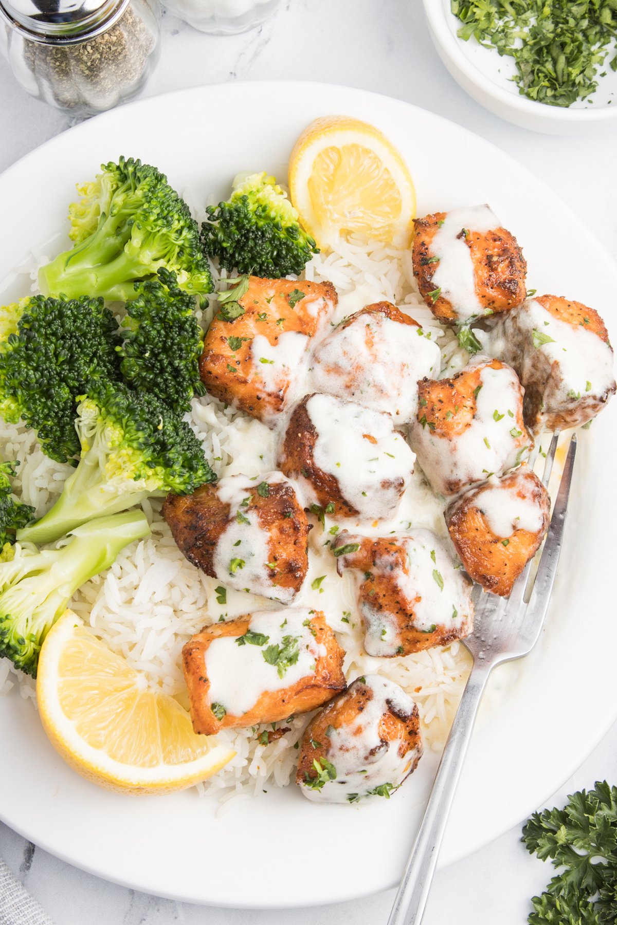 A white dinner plate with salmon bites covered in garlic cream sauce with white rice and broccoli. There are also two lemon slices, one has been squeezed.