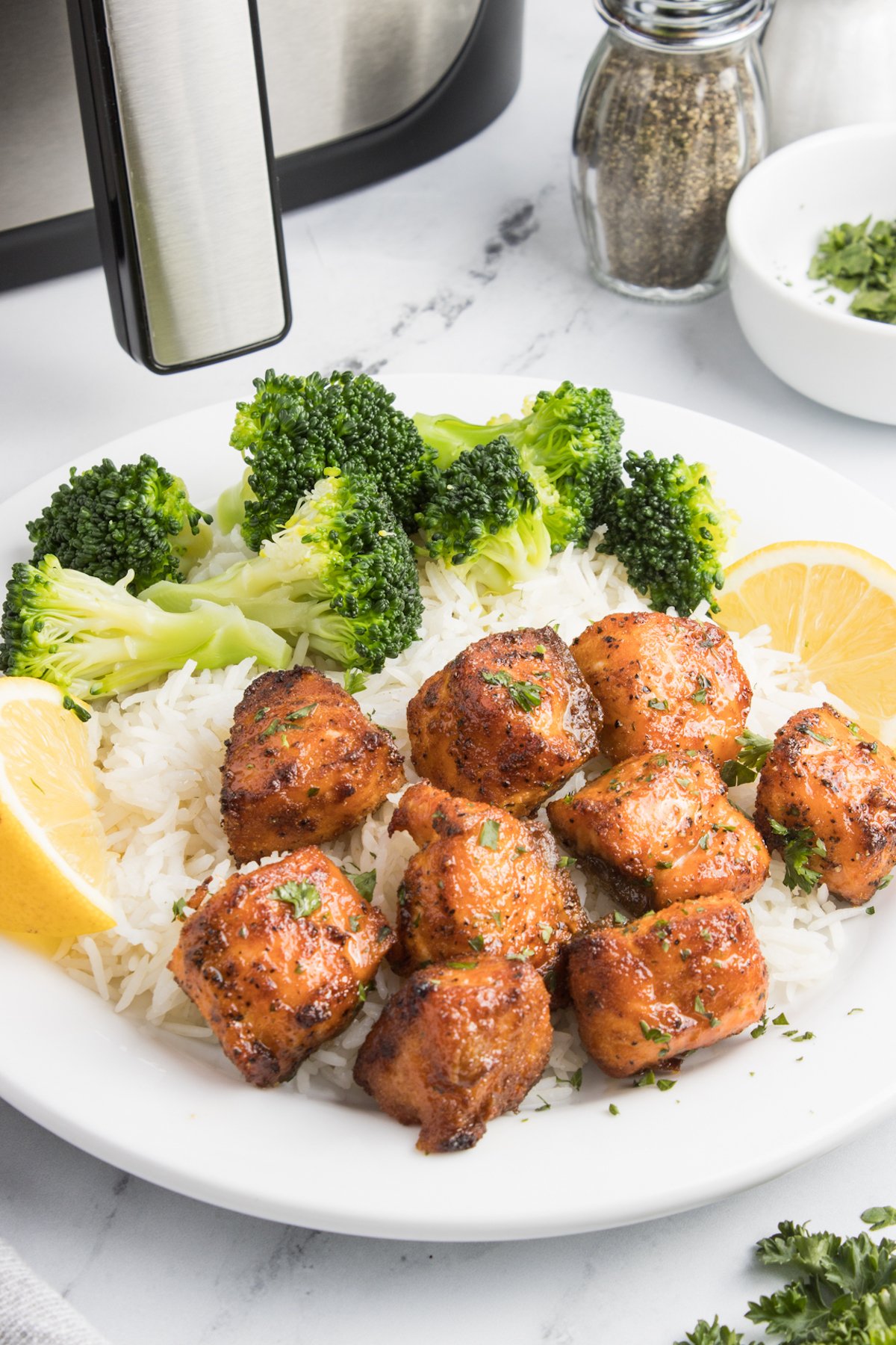 A white dinner plate with salmon bites, white rice, broccoli, and lemon slices in front of an air fryer.
