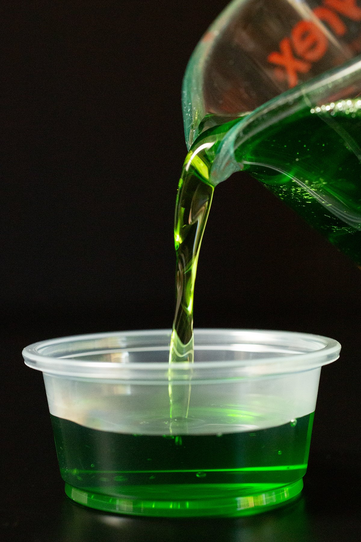 Liquid green apple gelatin and Crown mixture being poured into a 2 oz jello shot cup. 