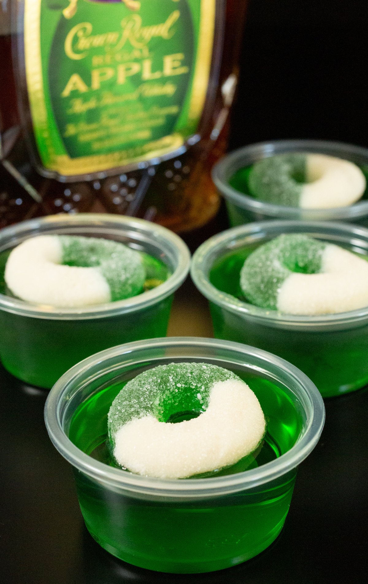 Four 2oz plastic containers filled with green crown apple jello shots topped with a green apple candy ring. 