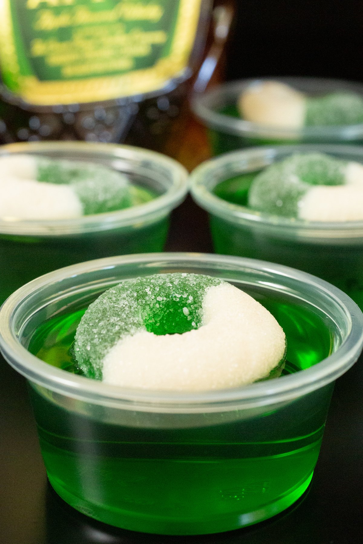 Close up of a 2oz plastic container filled with bright green Crown Apple jello shot that is topped with an apple candy ring.