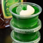 Four crown apple green jello shots stacked on top of each other. A bottle of Crown Apple is out of focus in the background.