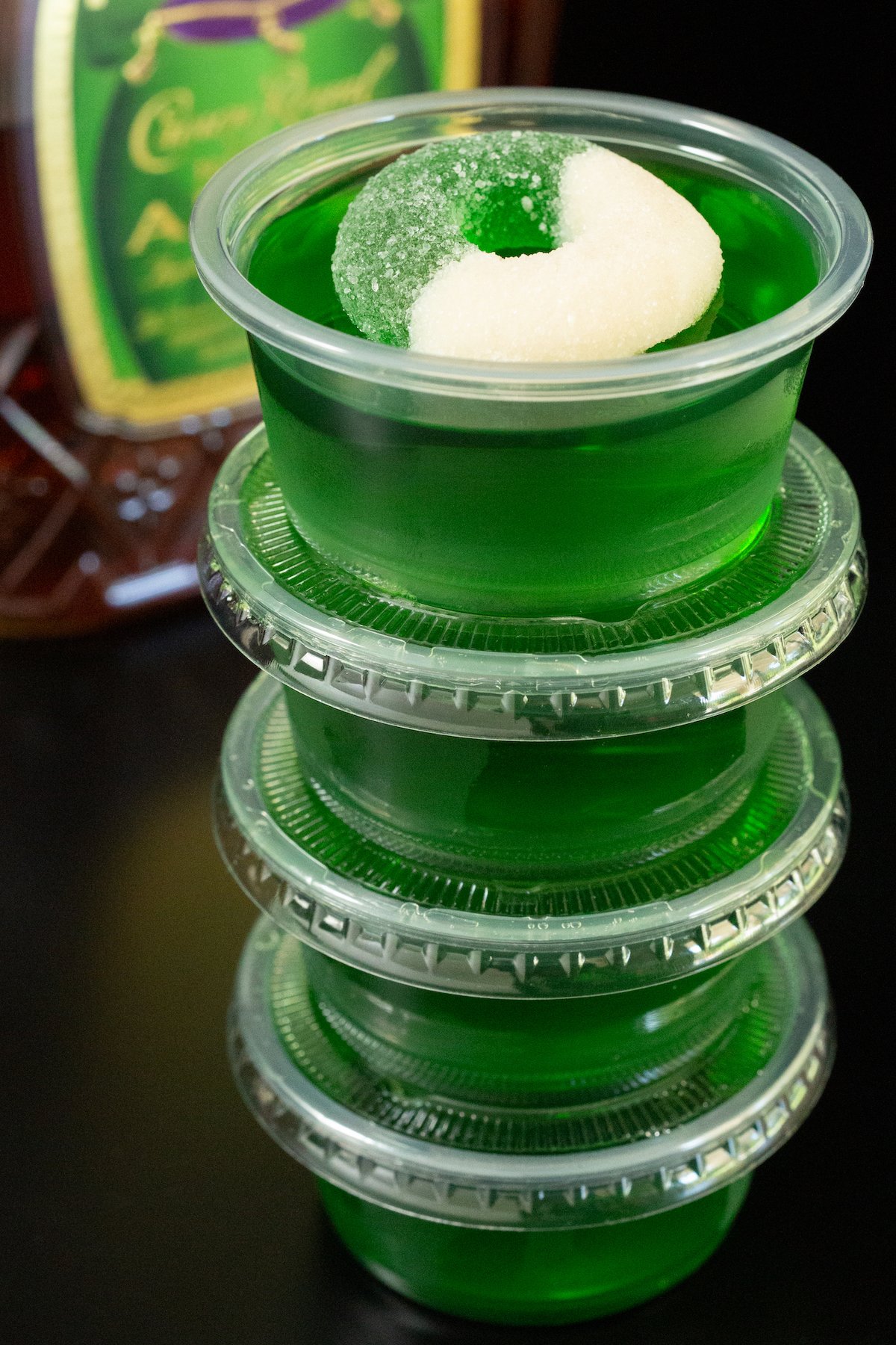 Four crown apple green jello shots stacked on top of each other. A bottle of Crown Apple is out of focus in the background.