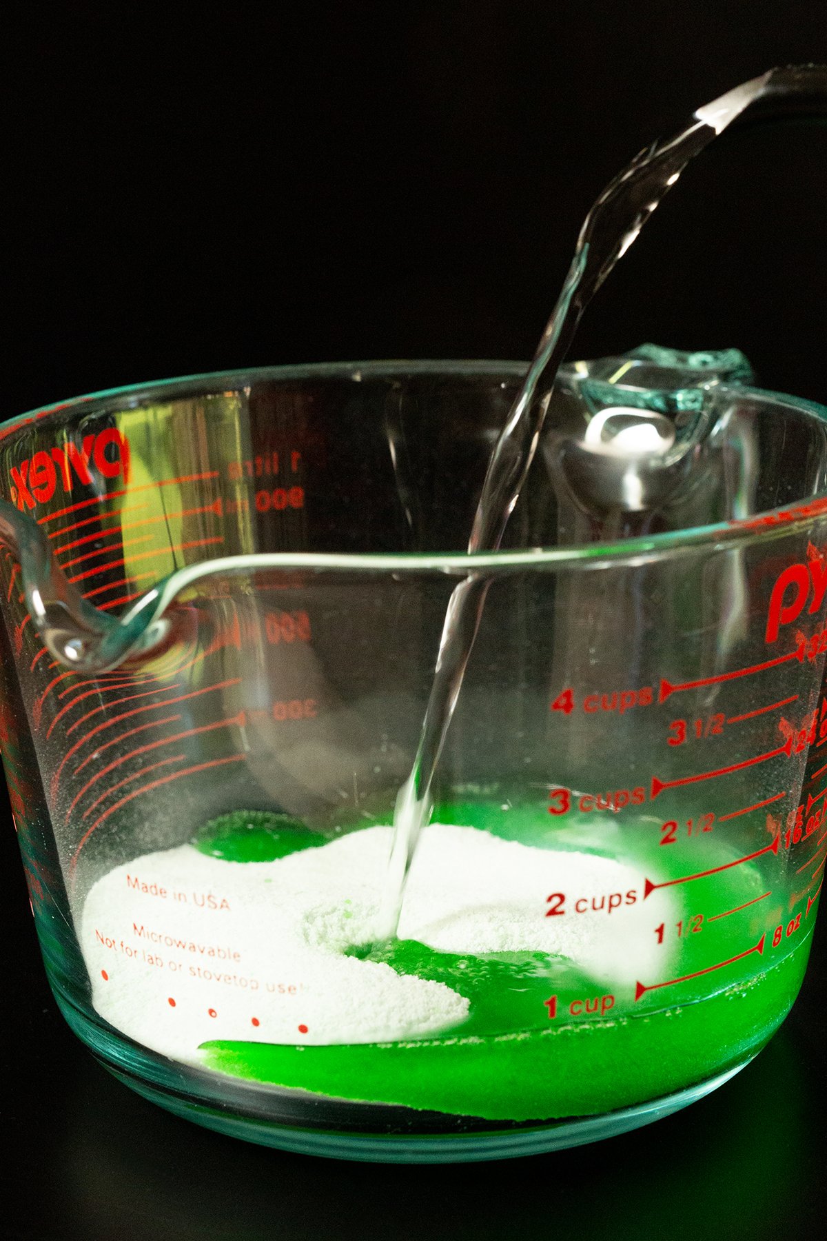 Hot water from a kettle being poured into a 4-cup glass measuring cup that has green apple gelatin powder in it.