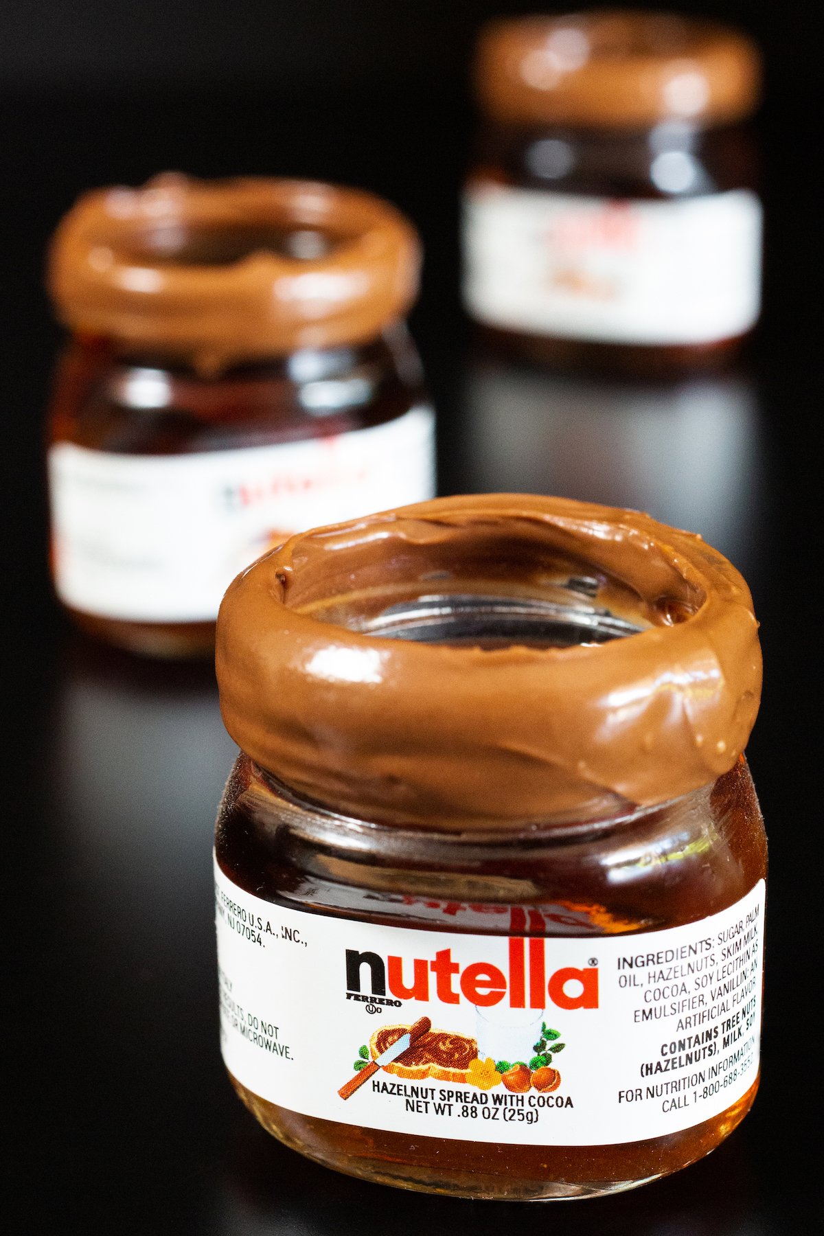 Three miniature Nutella jars filled with shots and rimmed with Nutella spread on a black background.