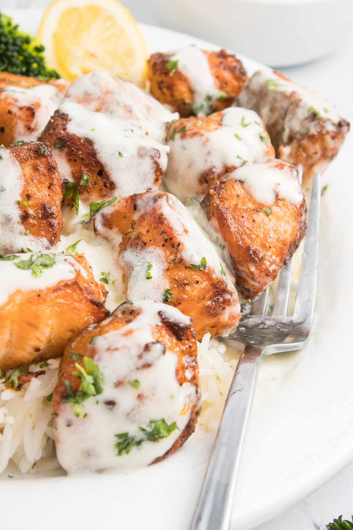 Close up of garlic salmon bites covered in garlic cream sauce next to a fork. A piece of broccoli and lemon slice are out of focus in the background.