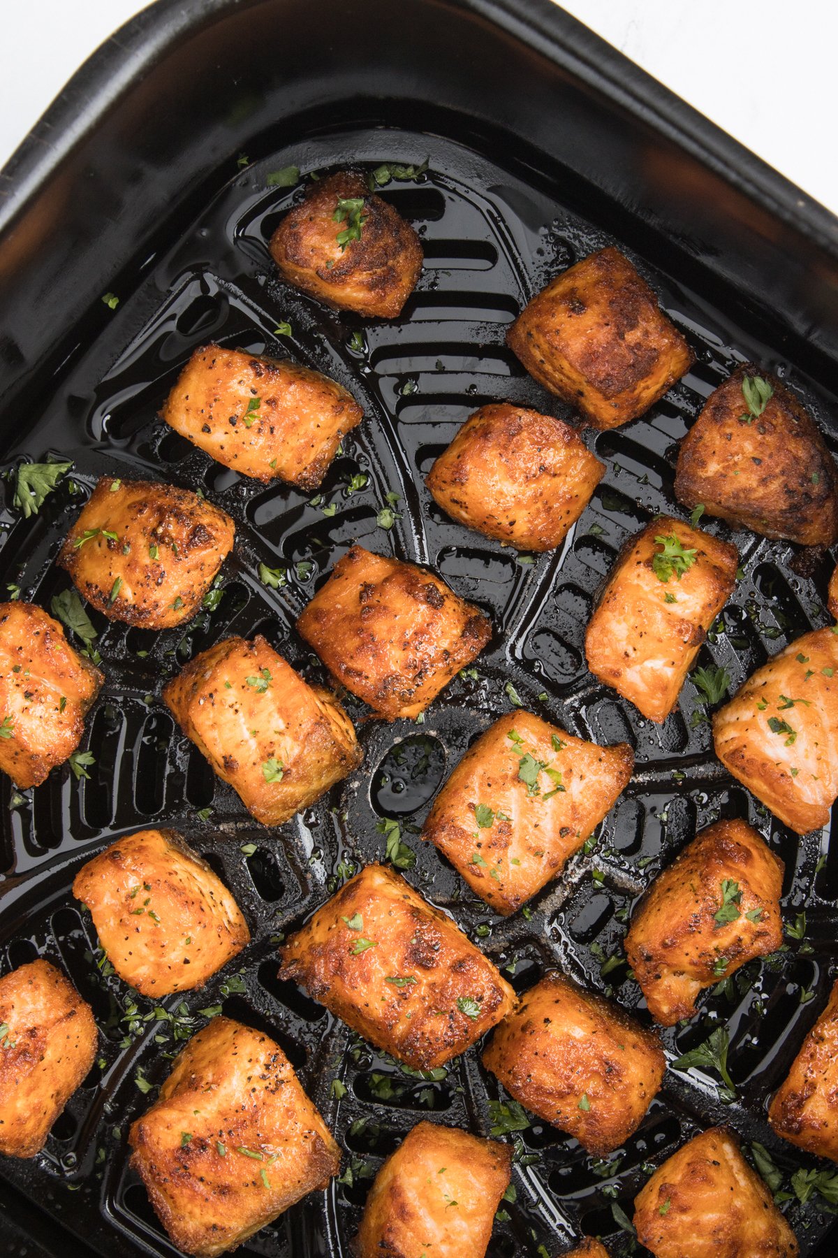 An air fryer basket filled with cooked salmon bites that are garnished with fresh chopped parsley.
