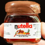 A thumb and index finger hold up a mini Nutella jar that's filled with a Nutella shot. The rim has been dipped in the hazelnut spread.