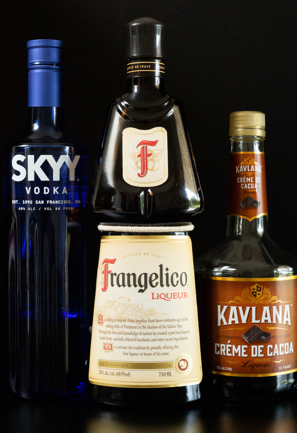 A bottle of Skyy vodka, Frangelico liqueur, and creme de cacao all on a black background.