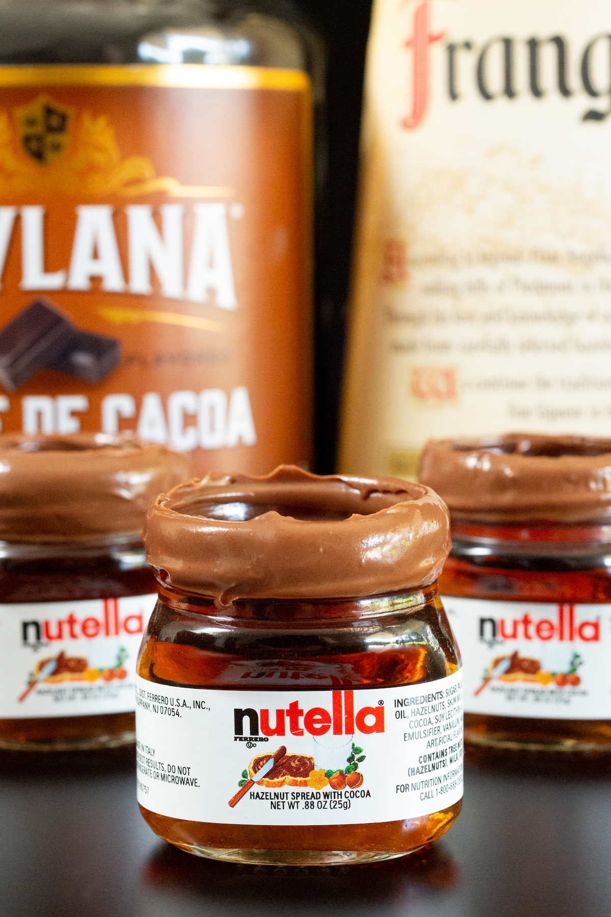 Three miniature Nutella jars with Nutella shots in them in front of creme de cacao and Fragelico liqueur bottles.