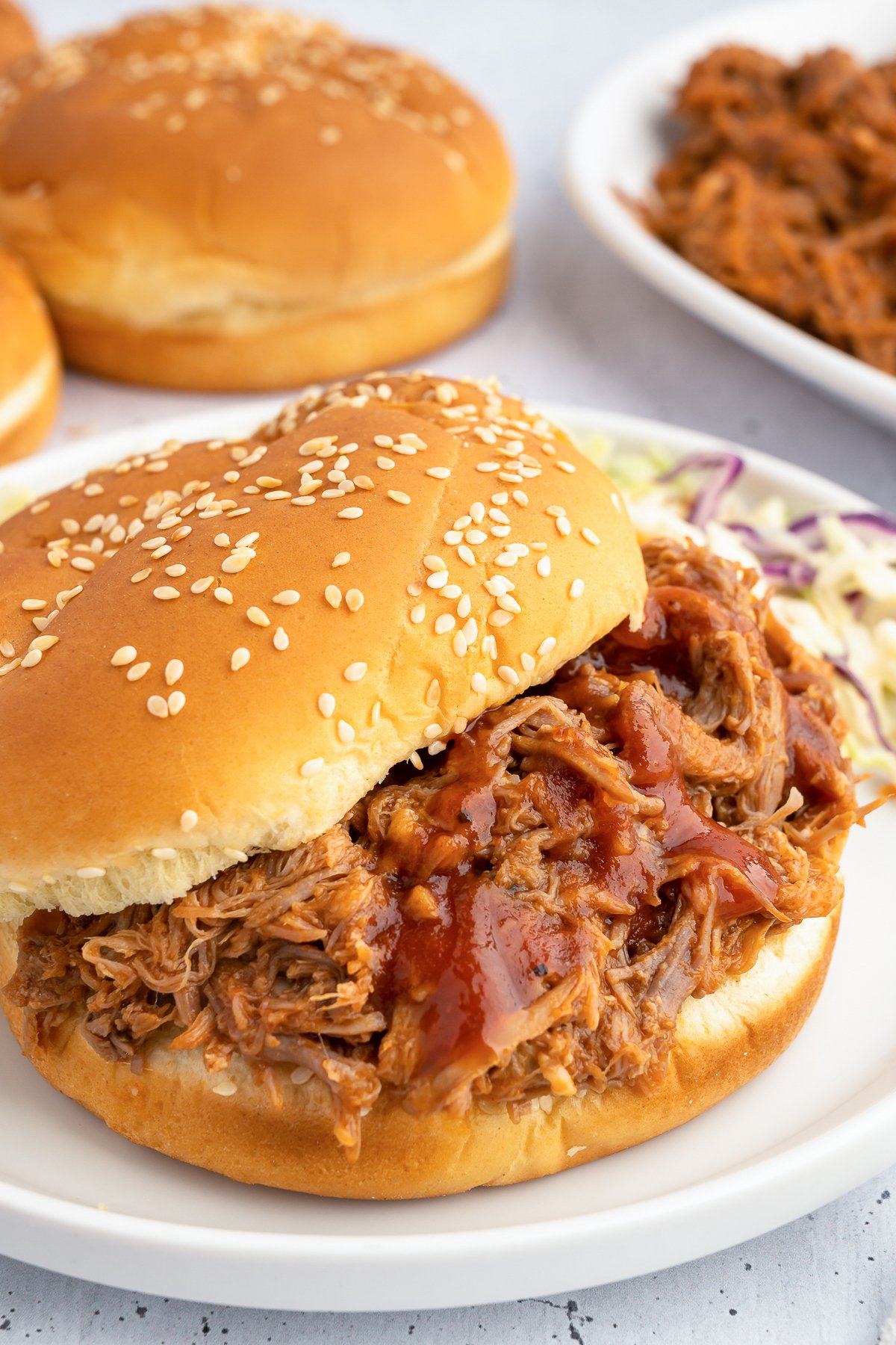 A pulled pork sandwich is in focus in the foreground, hamburger buns and more pork is out of focus in the background.