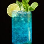 A tall glass is filled with pebble ice and a vibrant blue mojito that's garnished with a lime wheel and fresh mint.