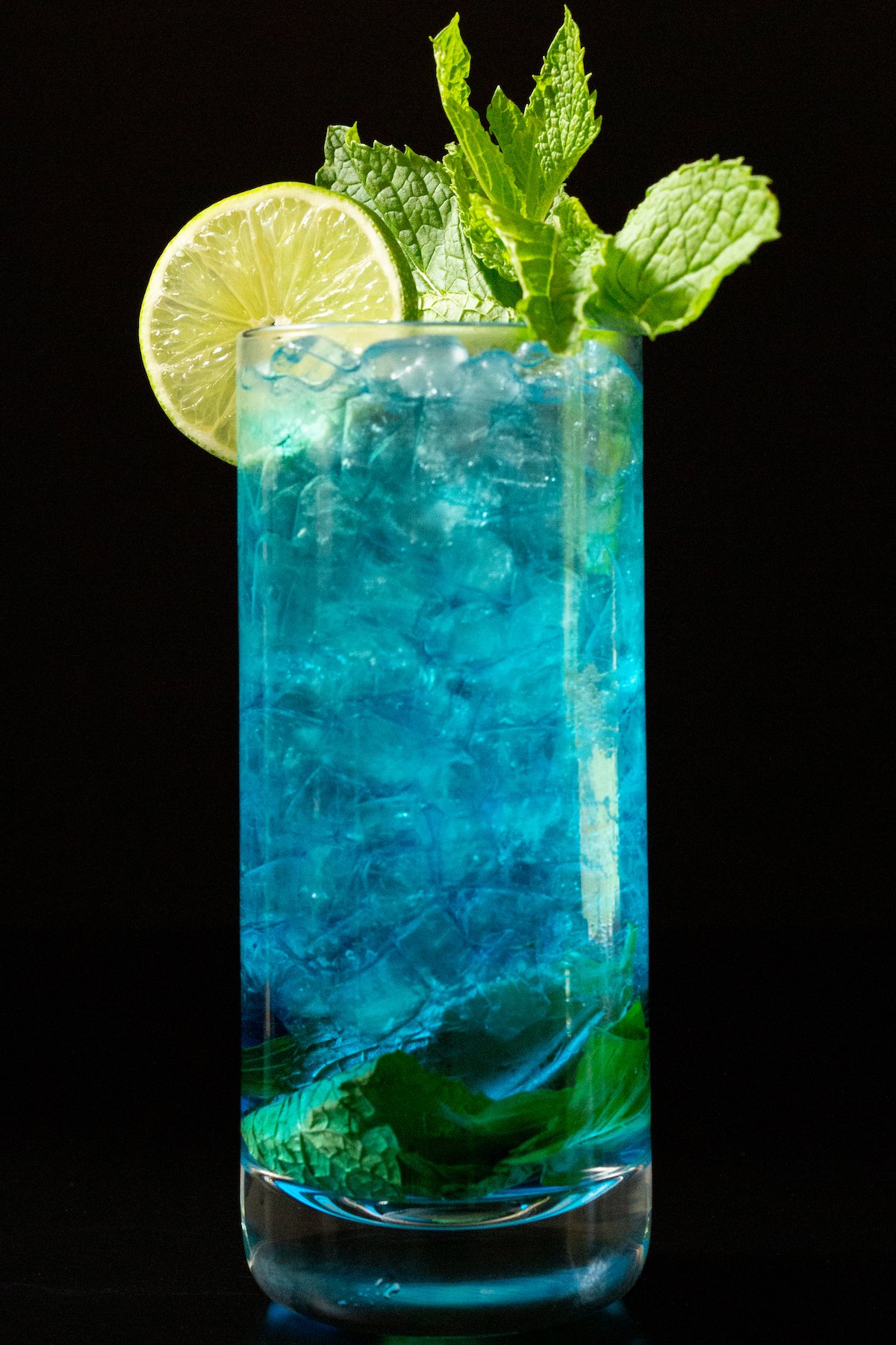 A tall glass is filled with pebble ice and a vibrant blue mojito that's garnished with a lime wheel and fresh mint.