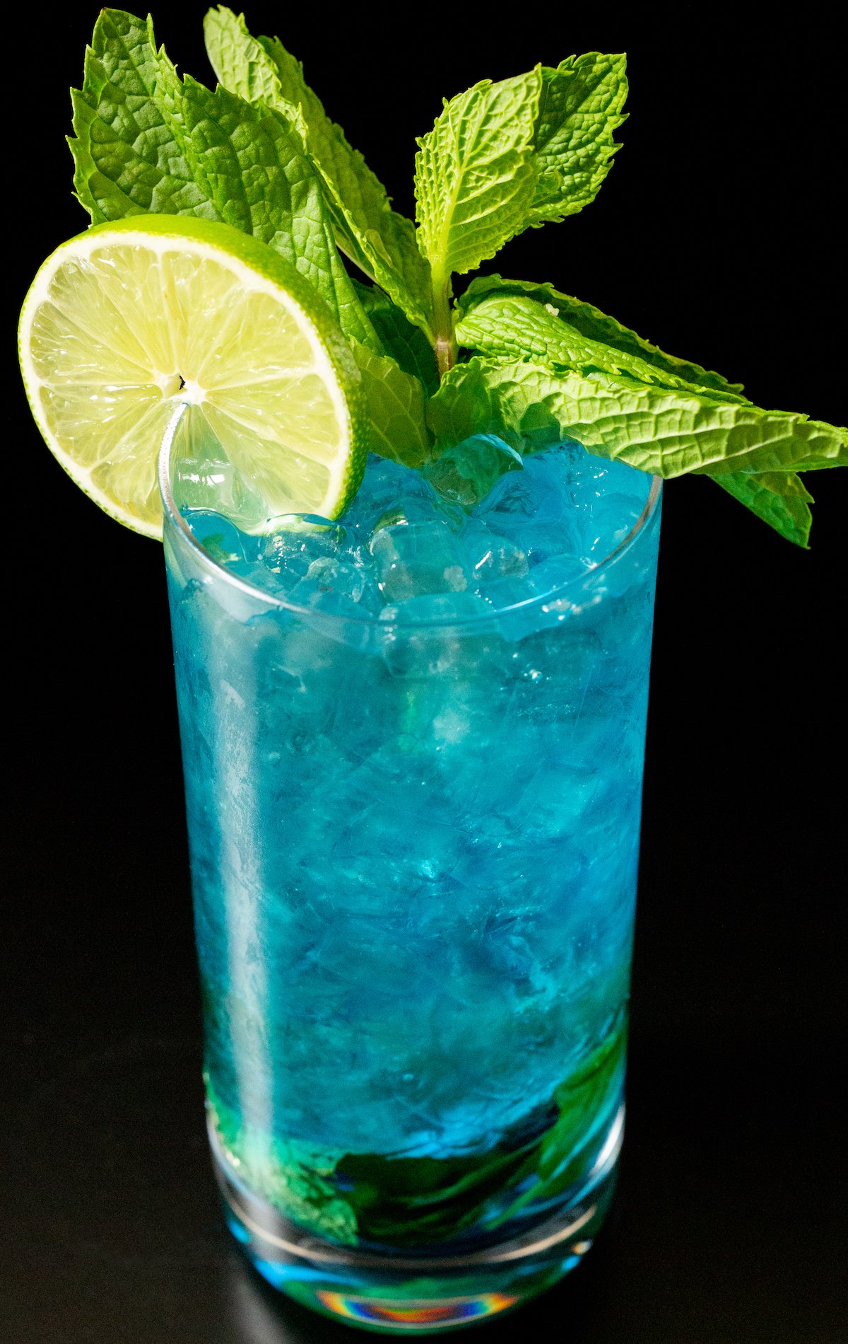 A tall glass filled with pebble ice and an ocean colored Blue Mojito that's garnished with a lime wheel and a mint sprig.