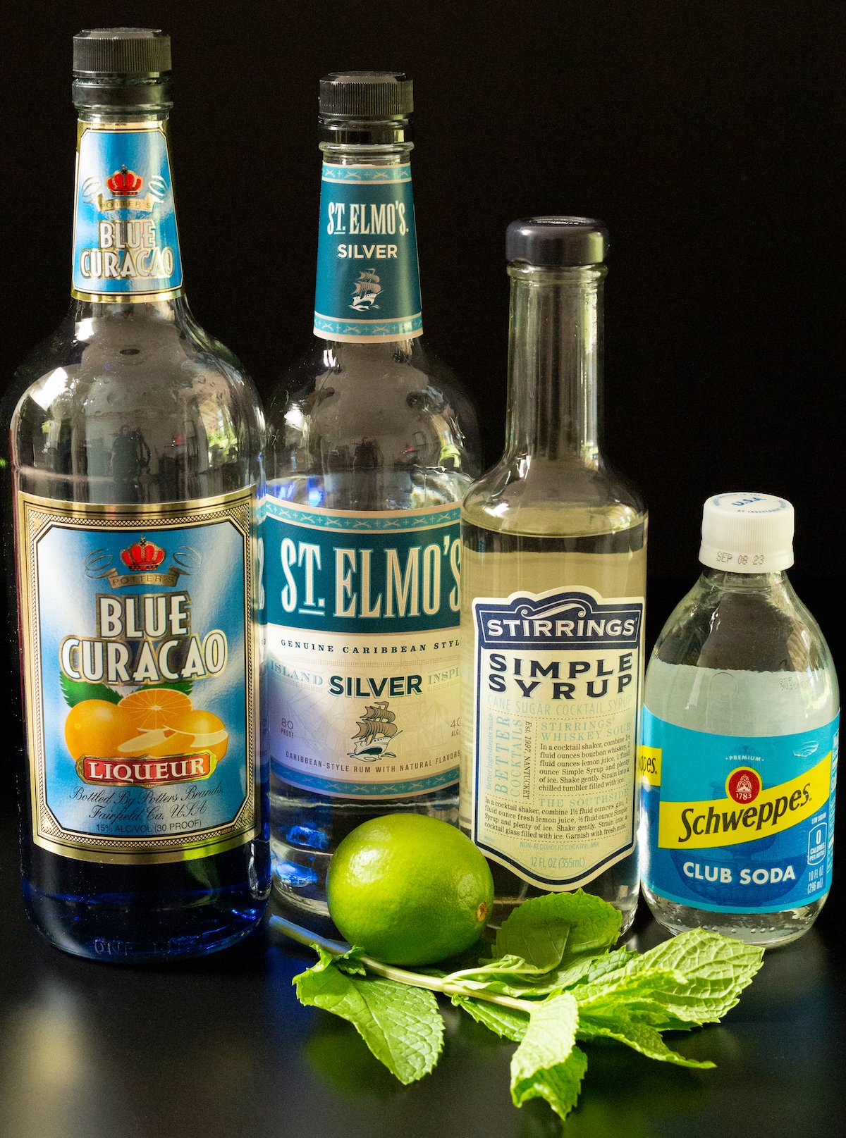 A bottle of blue curacao, white rum, simple syrup, club soda, a lime, and mint leaves on a black background.