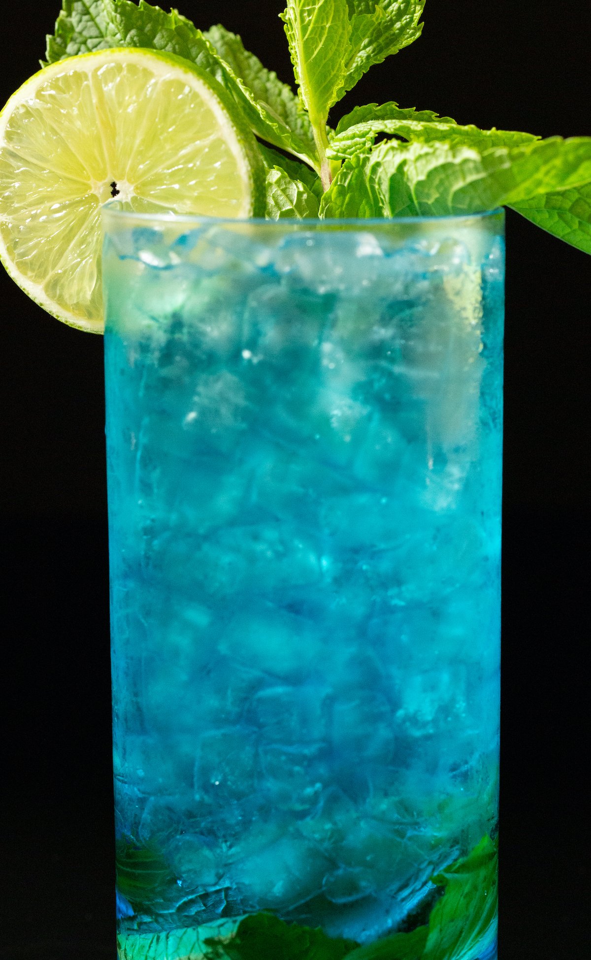 Close up of a blue mojito in a tall glass with a lime wheel and mint sprig garnish.