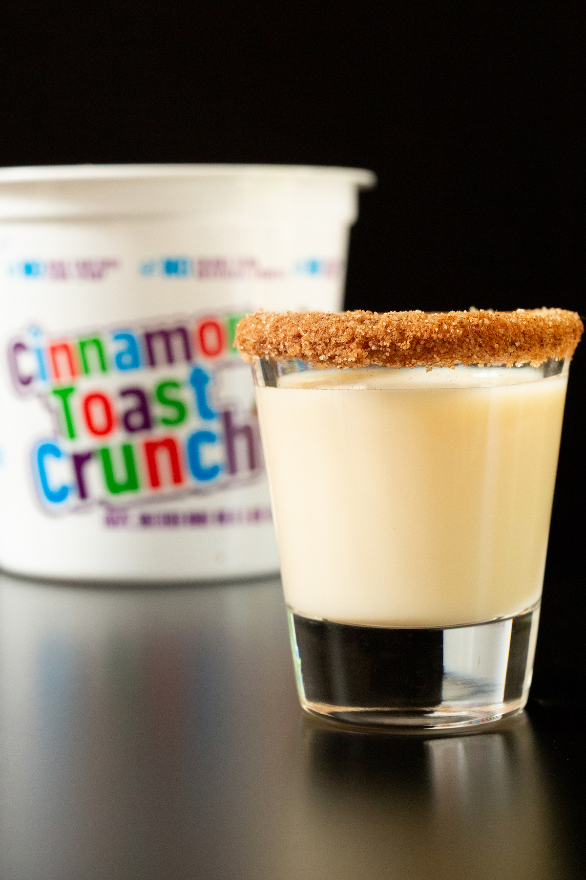 A single cinnamon toast crunch shot with cinnamon sugar rim in front of a container of the cereal.
