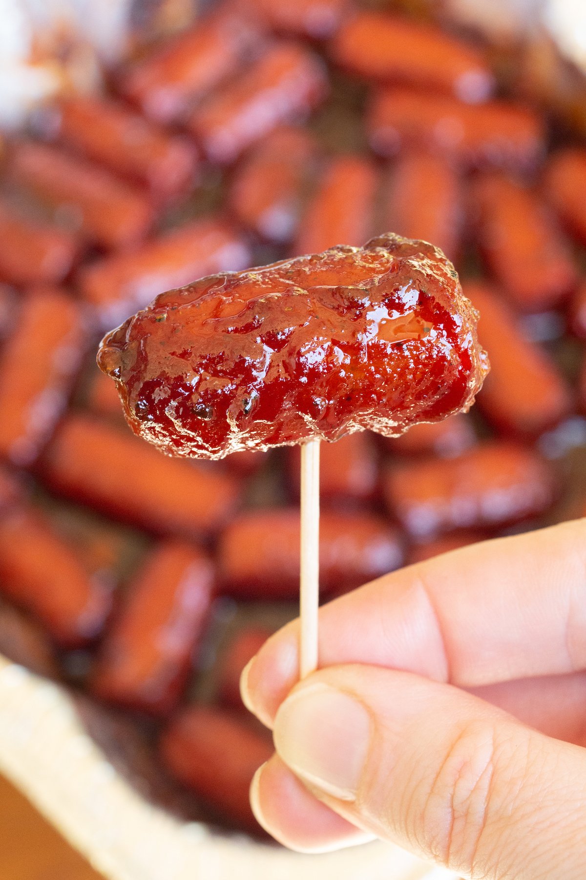 A hand holds up a single hot dog burnt end on a toothpick. A disposable pan with the rest is out of focus in the background.