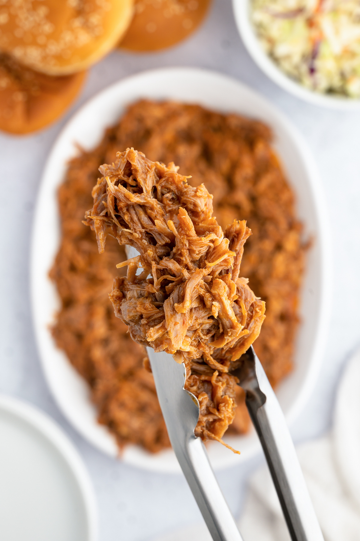 Tongs hold up Instant Pot pulled pork. A serving dish full of it is out of focus in the background.